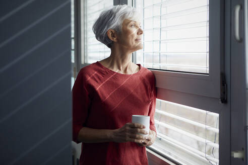 Porträt einer älteren Frau mit Kaffeetasse, die aus dem Fenster schaut - RBF07607
