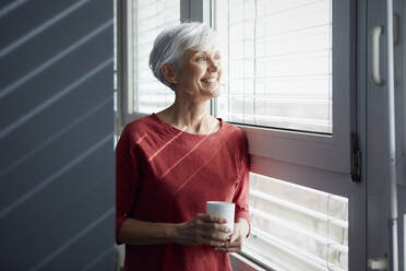 Porträt einer glücklichen älteren Frau mit Kaffeetasse, die aus dem Fenster schaut - RBF07608