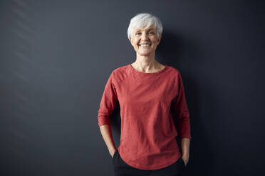 Porträt einer lächelnden älteren Frau mit rotem Hemd vor einer grauen Wand - RBF07609