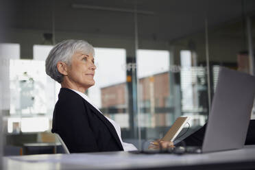 Ältere Geschäftsfrau sitzt am Schreibtisch im Büro und schaut in die Ferne - RBF07617