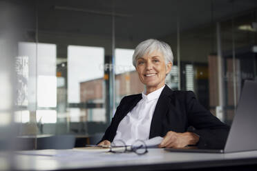 Porträt einer lächelnden älteren Geschäftsfrau, die am Schreibtisch im Büro sitzt - RBF07618