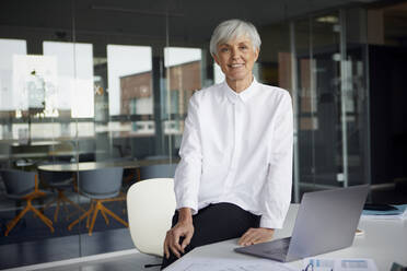 Portrait of smiling senior businesswoman sitting on desk in her office - RBF07626