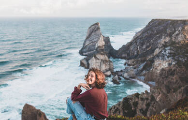 Smiling young woman sitting on viewpoint at Praia da Ursa, Lisboa, Portugal - FVSF00179