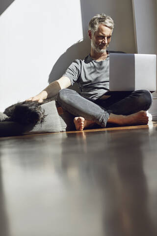 Älterer Mann sitzt zu Hause auf dem Boden und schaut auf seinen Laptop, während er seinen Hund streichelt, lizenzfreies Stockfoto