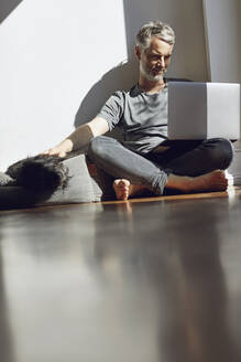 Mature man sitting on the floor at home looking at laptop while stroking his dog - MCF00775