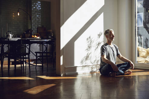 Mature man sitting on the floor at home meditating - MCF00776