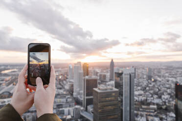 Deutschland, Hessen, Frankfurt, Hände einer Frau, die mit ihrem Smartphone Fotos von der Innenstadt bei Sonnenuntergang macht - AHSF02325