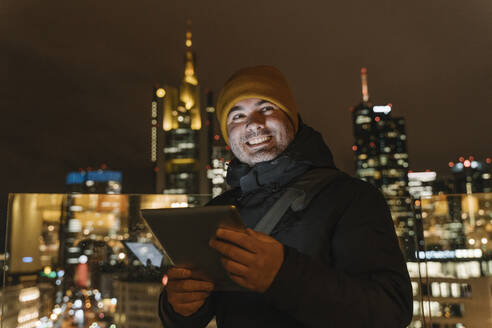 Deutschland, Hessen, Frankfurt, Porträt eines erwachsenen Mannes, der nachts im Freien steht und mit einem digitalen Tablet in der Hand lächelt - AHSF02338