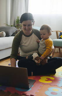 Mutter und ihr kleiner Sohn sitzen auf dem Boden des Wohnzimmers und schauen auf den Laptop - BZF00552
