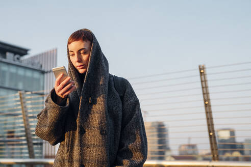 Rothaarige junge Frau mit Smartphone auf einer Brücke in der Stadt - MEUF00469