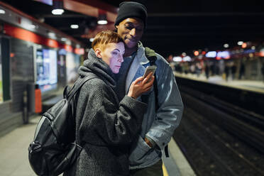 Ein junges Paar teilt sich ein Smartphone auf dem Bahnsteig - MEUF00515