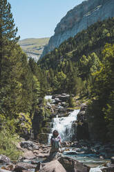 Spain, Province of Huesca, Female hiker admiring small waterfall on clear mountain river - FVSF00217