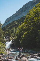 Spain, Province of Huesca, Female hiker raising arms while admiring small waterfall on clear mountain river - FVSF00218