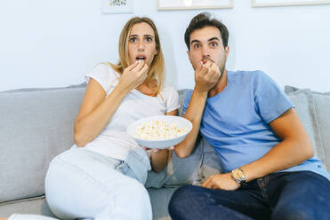 Aufgeregtes Paar beim Fernsehen und Popcorn essen auf dem Sofa zu Hause - KIJF02981