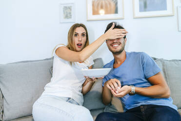 Shocked woman covering man's eyes while watching TV in living room at home - KIJF02982