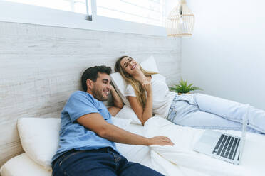 Glückliches Paar sieht sich einen Film auf dem Laptop an, während es zu Hause auf dem Bett liegt - KIJF02989
