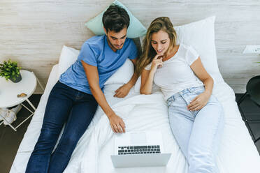 Blick von oben auf ein Paar, das einen Film auf dem Laptop ansieht, während es sich zu Hause auf dem Bett entspannt - KIJF02990