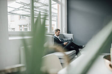 Geschäftsmann mit Smartphone auf der Couch im Büro sitzend - GUSF03502