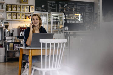 Lächelnde junge Frau mit Smartphone und Laptop in einem Café - GUSF03513