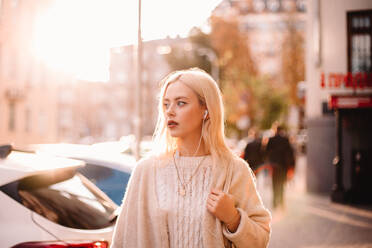 Young woman listening music on earphones while walking on city street - CAVF79269