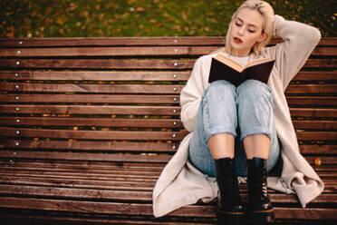 Young woman reading book sitting on bench in park during autumn - CAVF79282