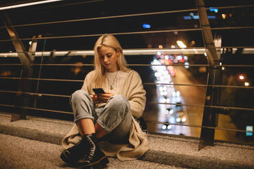 Teenager-Mädchen mit Smartphone sitzt auf einer Brücke in der Stadt bei Nacht - CAVF79289