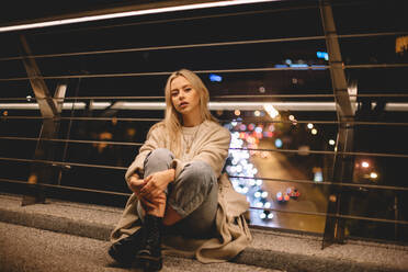 Portrait of young woman sitting on bridge in city at night - CAVF79290