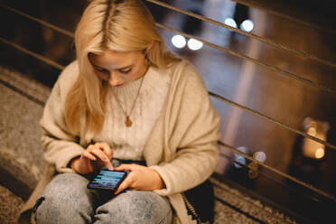 Junge Frau benutzt ihr Smartphone, während sie nachts auf einer Brücke in der Stadt sitzt - CAVF79291