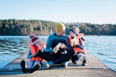 Ein Vater sitzt im Neoprenanzug auf einem Steg und isst mit seinen Kindern am Strand Obst - CAVF79381