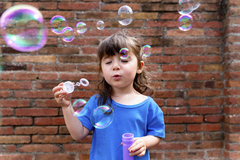 Portrait of little girl with eyes closed blowing soap bubbles in front of brick wall - GEMF03603