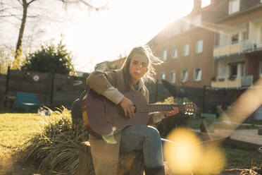 Junge Frau spielt Gitarre im Garten bei Sonnenschein - GUSF03551