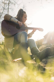 Junge Frau spielt Gitarre im Garten bei Sonnenschein - GUSF03553