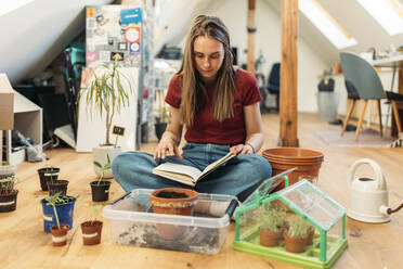 Junge Frau liest ein Buch neben Pflanzen auf einem Holzboden - GUSF03563