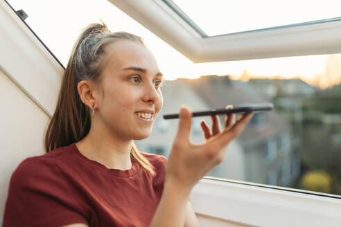 Junge Frau mit Smartphone am Fenster im Gegenlicht - GUSF03590