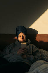 Young woman with head wrapped in a towel lying in bed at home using smartphone - GUSF03642