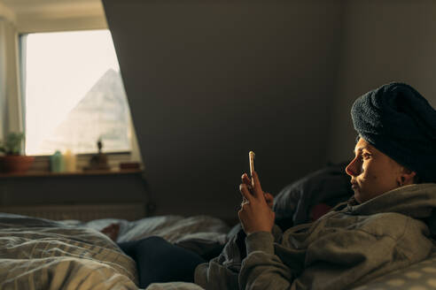 Junge Frau mit in ein Handtuch gewickeltem Kopf, die zu Hause im Bett liegt und ihr Smartphone benutzt - GUSF03644