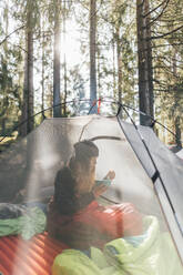 Junge Frau sitzt im Zelt und frühstückt im Wald - GUSF03694