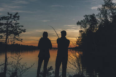 Couple standing at lake, fishing in lake at sunset - GUSF03700