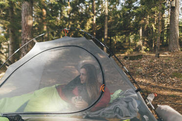 Young woman in tent isn the forest, drinking tea - GUSF03703