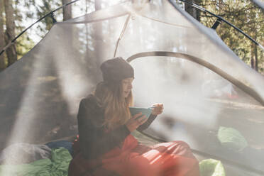 Junge Frau sitzt im Zelt und frühstückt im Wald - GUSF03706