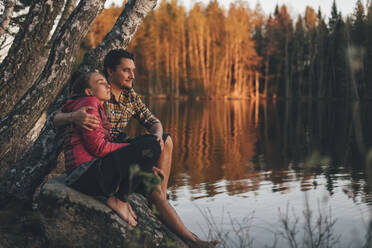 Junges Paar sitzt auf einem Stein am Seeufer und betrachtet den Sonnenuntergang - GUSF03726