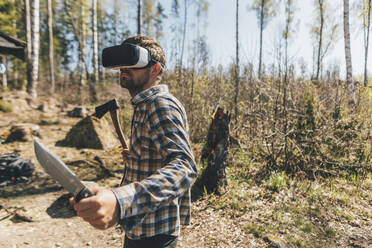 Young man gaming with VR glasses in the forest, holding axe and knife - GUSF03738