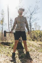 Young man gaming with VR glasses in the forest, holding axe - GUSF03739