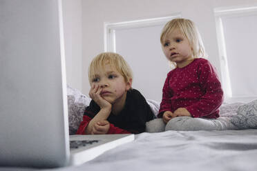 Porträt eines kleinen Mädchens und ihres älteren Bruders auf dem Bett mit Blick auf den Laptop - JOSEF00319