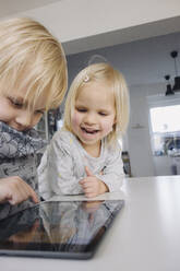 Porträt eines lachenden kleinen Mädchens, das seinen älteren Bruder bei der Benutzung eines digitalen Tablets beobachtet - JOSEF00324