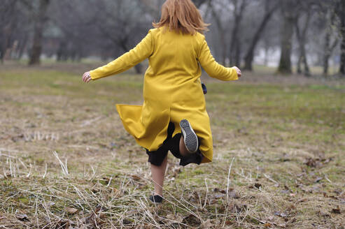 Rückansicht einer Frau im gelben Mantel, die im Park auf dem Rasen läuft - EYAF01042
