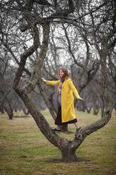 Frau in gelbem Mantel in voller Länge auf einem kahlen Baum im Park stehend - EYAF01044