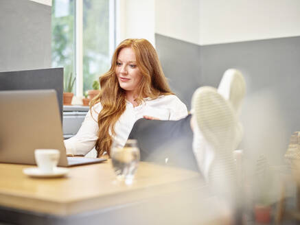 Geschäftsfrau in voller Länge, die einen Laptop benutzt, während sie sich in der Kaffeepause am Tisch entspannt - CVF01623