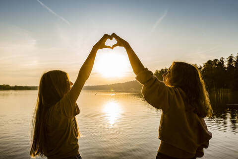 Freunde machen Herzform mit Händen gegen See bei Sonnenuntergang, lizenzfreies Stockfoto