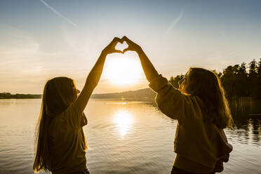Friends making heart shape with hands against lake during sunset - OJF00393